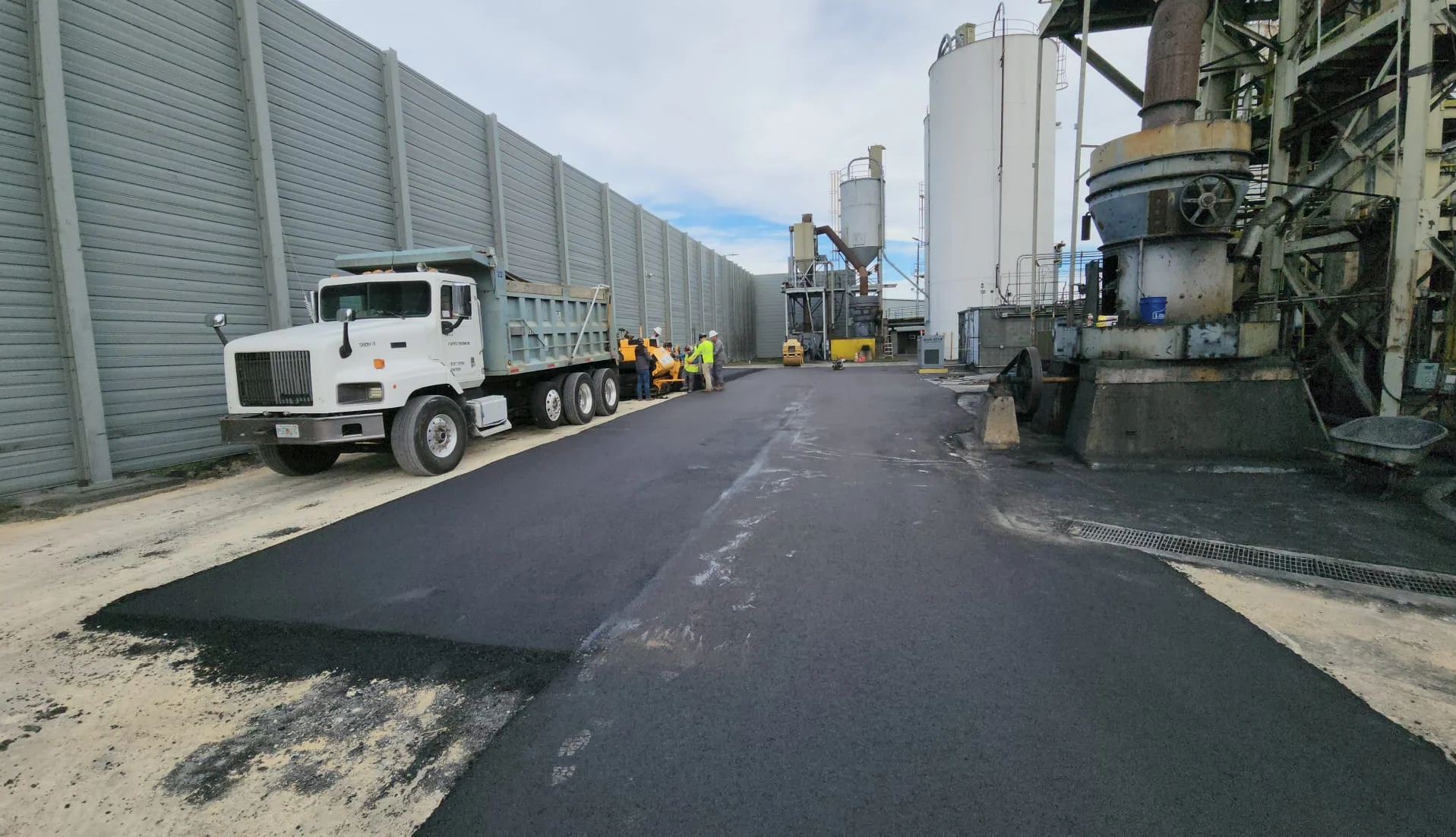A white dump truck next to workers using an asphalt paver to lay blacktop at an industrial facility.